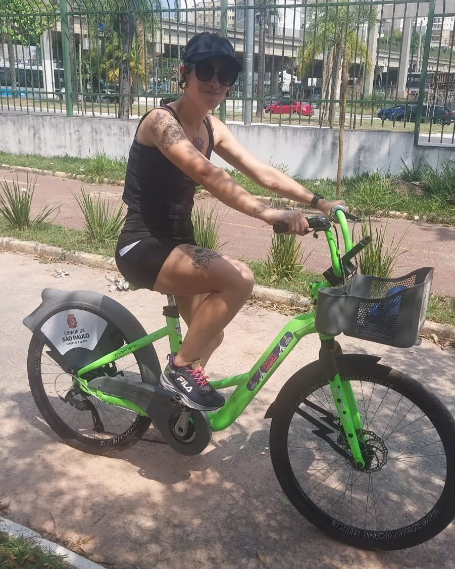 eu - mulher branca, tatuada,cabelos castanhos, vestindo shorts de academia preto, regata preta, tênis de corrida, óculos escuros e uma viseira preta - em cima de uma bicicleta verde com o logo da prefeitura de são paulo, em um parque com a avenida por trás, em um dia ensolarado